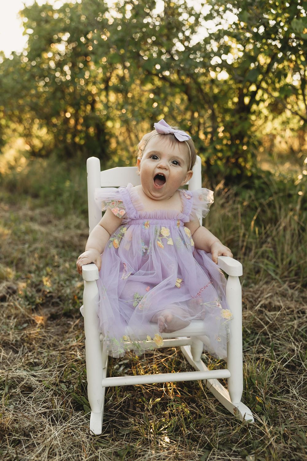 Baby girl in purple tulle dress sits in white rocking chair outdoors, mouth open wide, wearing lavender bow headband.