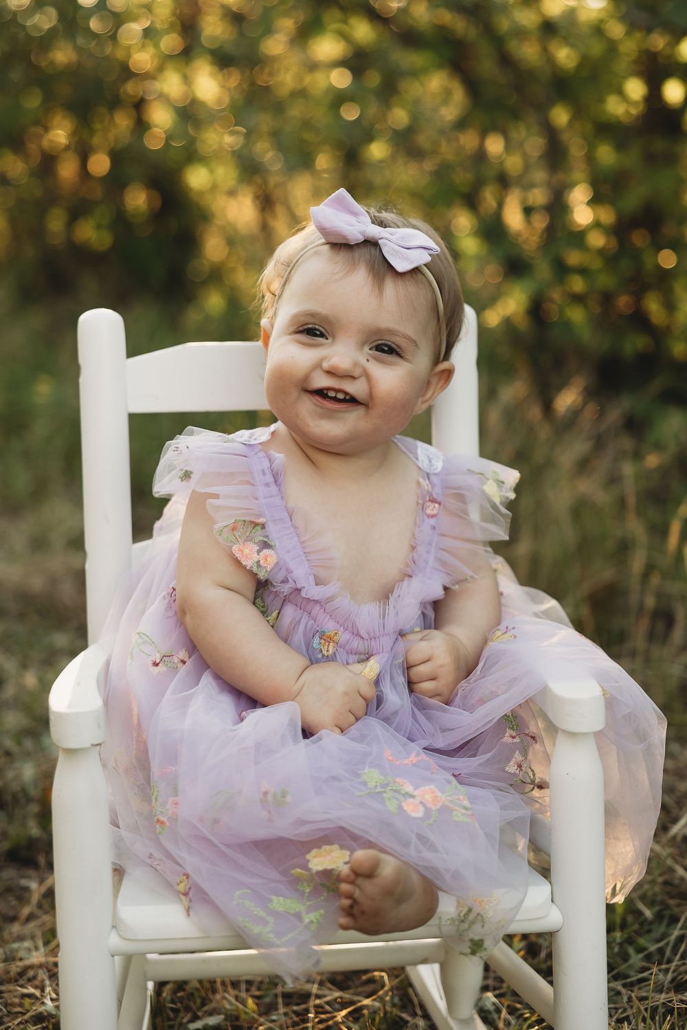Smiling baby girl in lavender tulle dress and bow headband sitting on white chair outdoors.