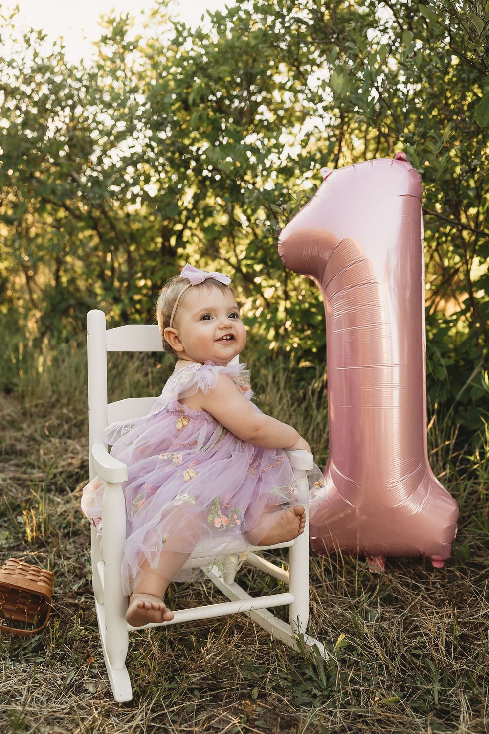 A baby girl in a pink tulle dress sits on a white chair outdoors next to a large pink number 1 balloon.