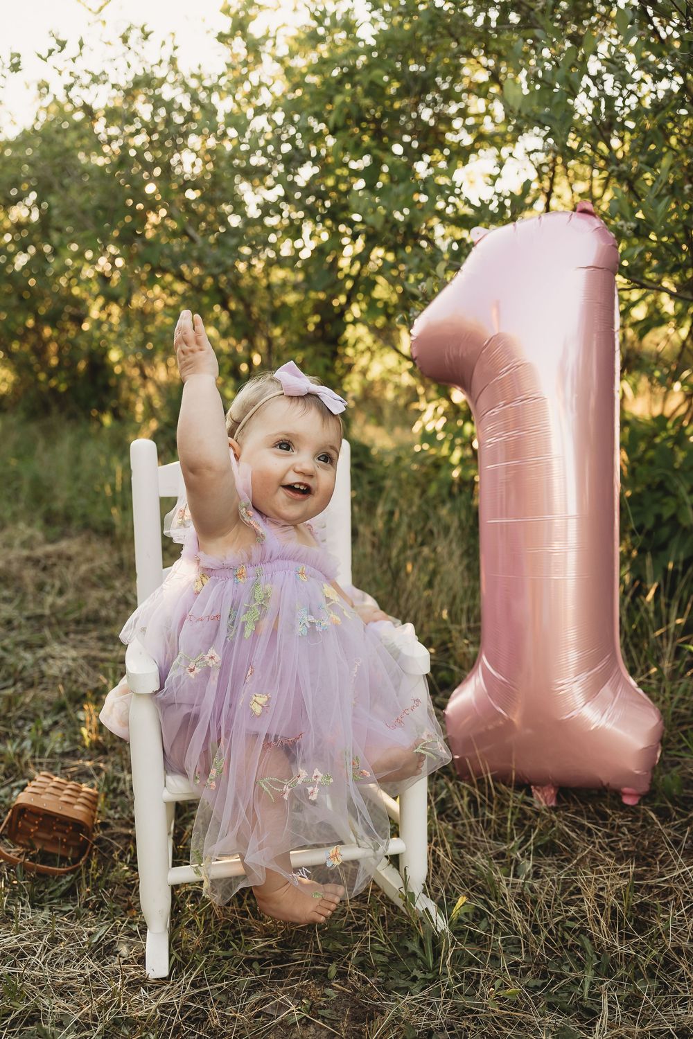Baby girl in purple tutu dress celebrates 1st birthday outdoors with pink number one balloon on white chair.
