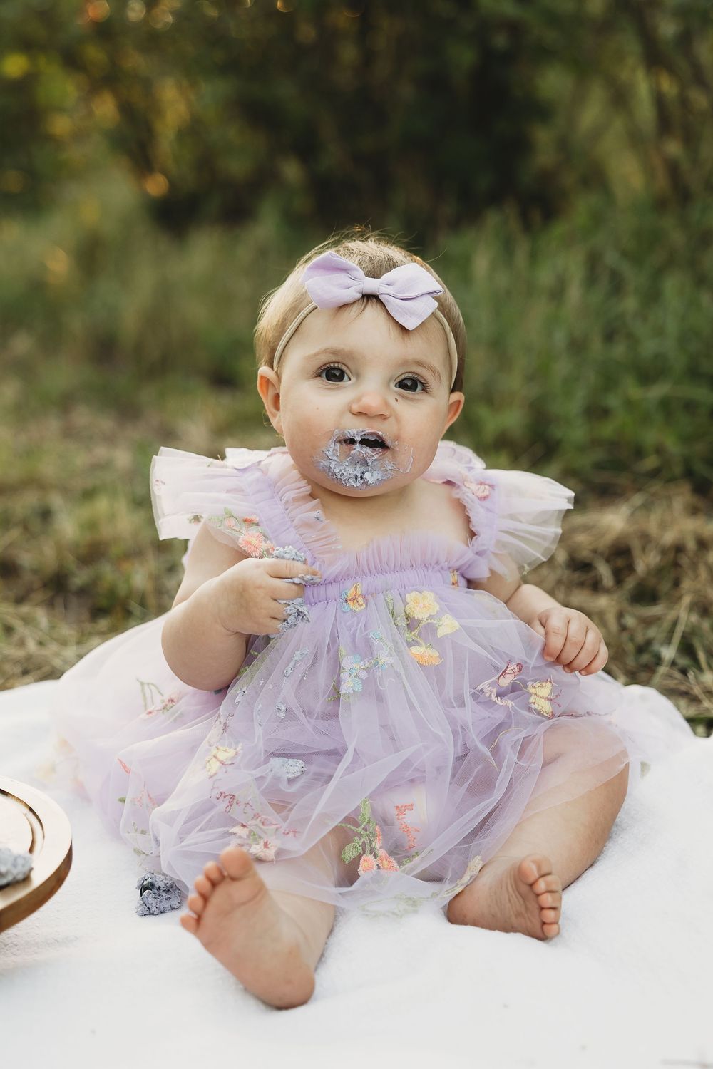 Baby girl in purple tulle dress with bow headband sits on white blanket outdoors, cake smash frosting on her face.