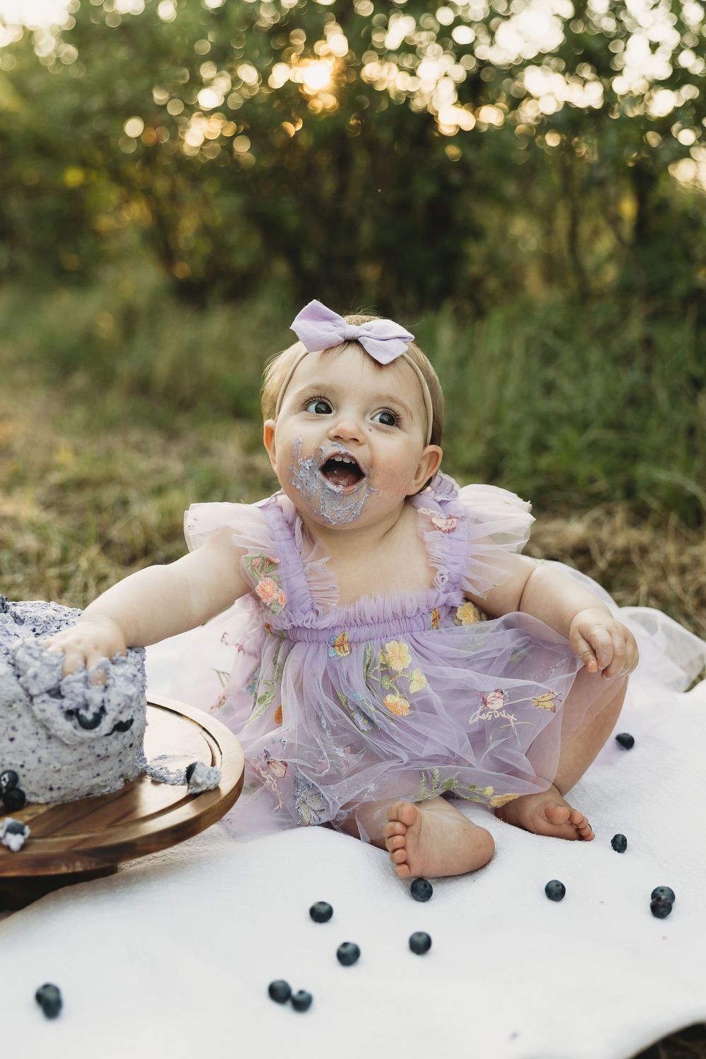 Baby girl in purple tulle dress enjoys first birthday cake smash outdoors with blueberries scattered around.
