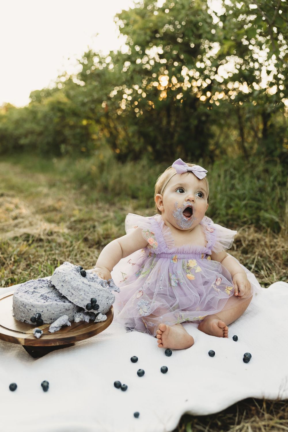 Baby girl in purple tutu dress eating blueberry smash cake outdoors during golden hour first birthday celebration.