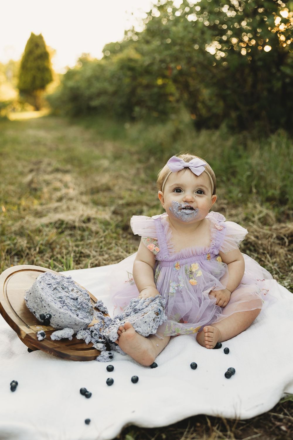 Baby girl in purple tutu sits outdoors with smashed blueberry cake on white blanket during golden hour birthday celebration.