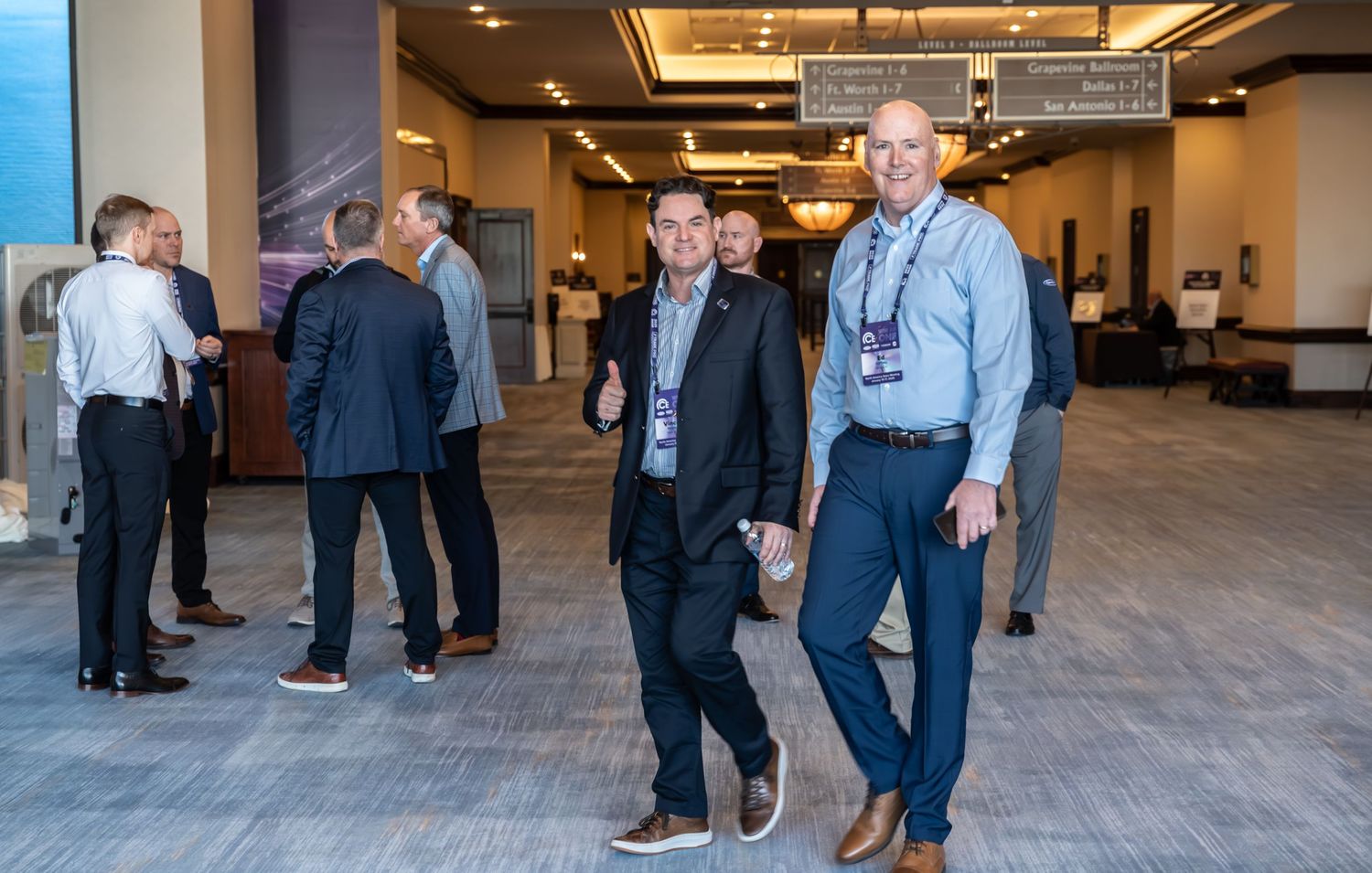 Two professionals in business attire smile and walk through a modern conference hall with attendees networking in the background.