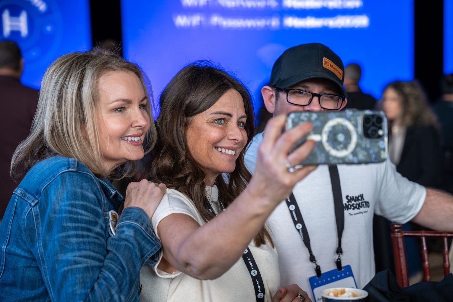 Three conference attendees smiling and taking a selfie at a professional networking event with blue presentation screens in background.