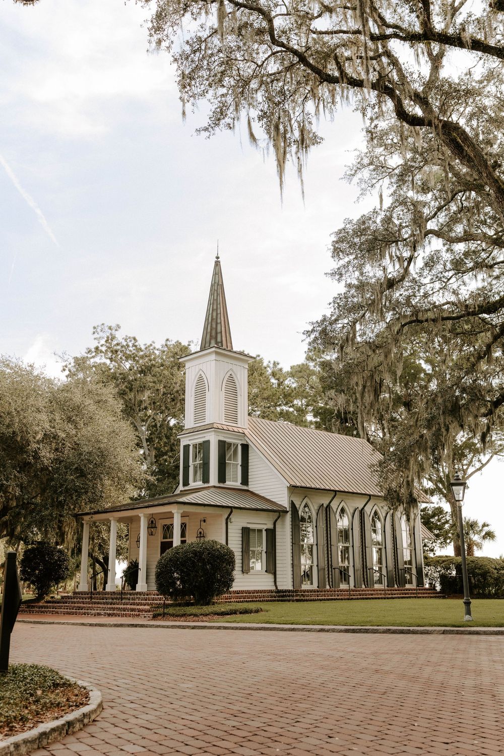 White Southern Gothic church with steeple, brick pathway, and Spanish moss-draped oak trees in a serene landscape.
