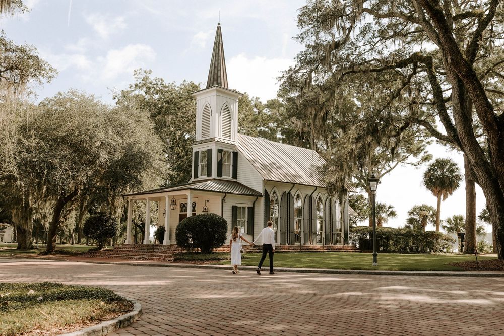 Couple walking in front of a white historic chapel surrounded by oak trees on a sunny day.