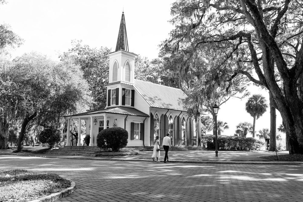 Black and white photo of a historic white church with steeple surrounded by large oak trees and brick pathways.