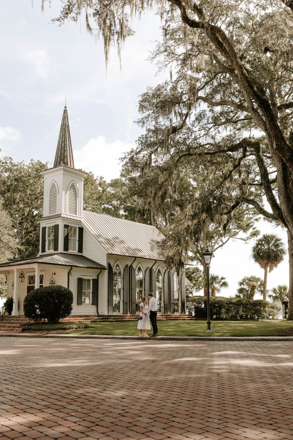 Couple standing before a white Gothic Revival chapel with Spanish moss-draped oak trees on a brick plaza.