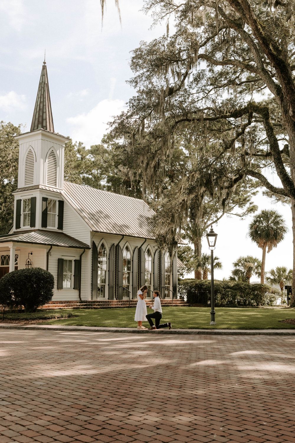 Man proposing on one knee to woman in white dress outside a white chapel with Spanish moss trees.