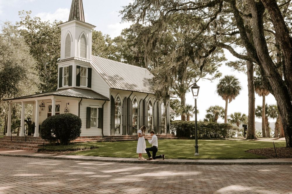 Couple sharing a romantic proposal moment outside a white chapel surrounded by oak trees draped in Spanish moss.