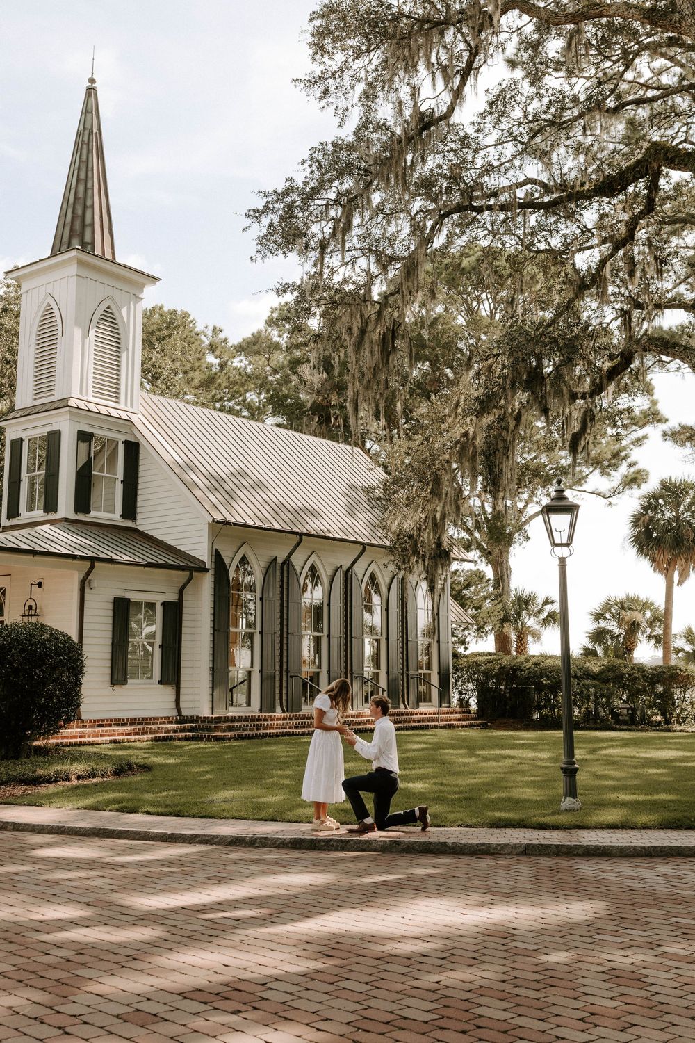 Man proposing on one knee to woman in white dress outside a white chapel with Spanish moss trees.