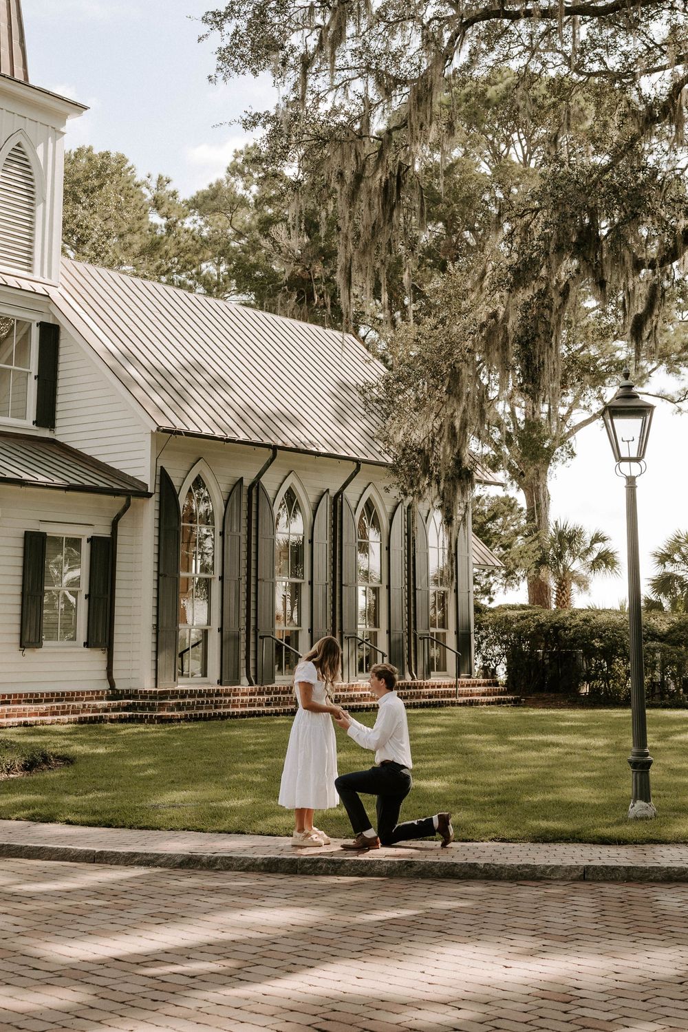 Man proposing on one knee to woman in white dress outside a white church with Spanish moss trees.