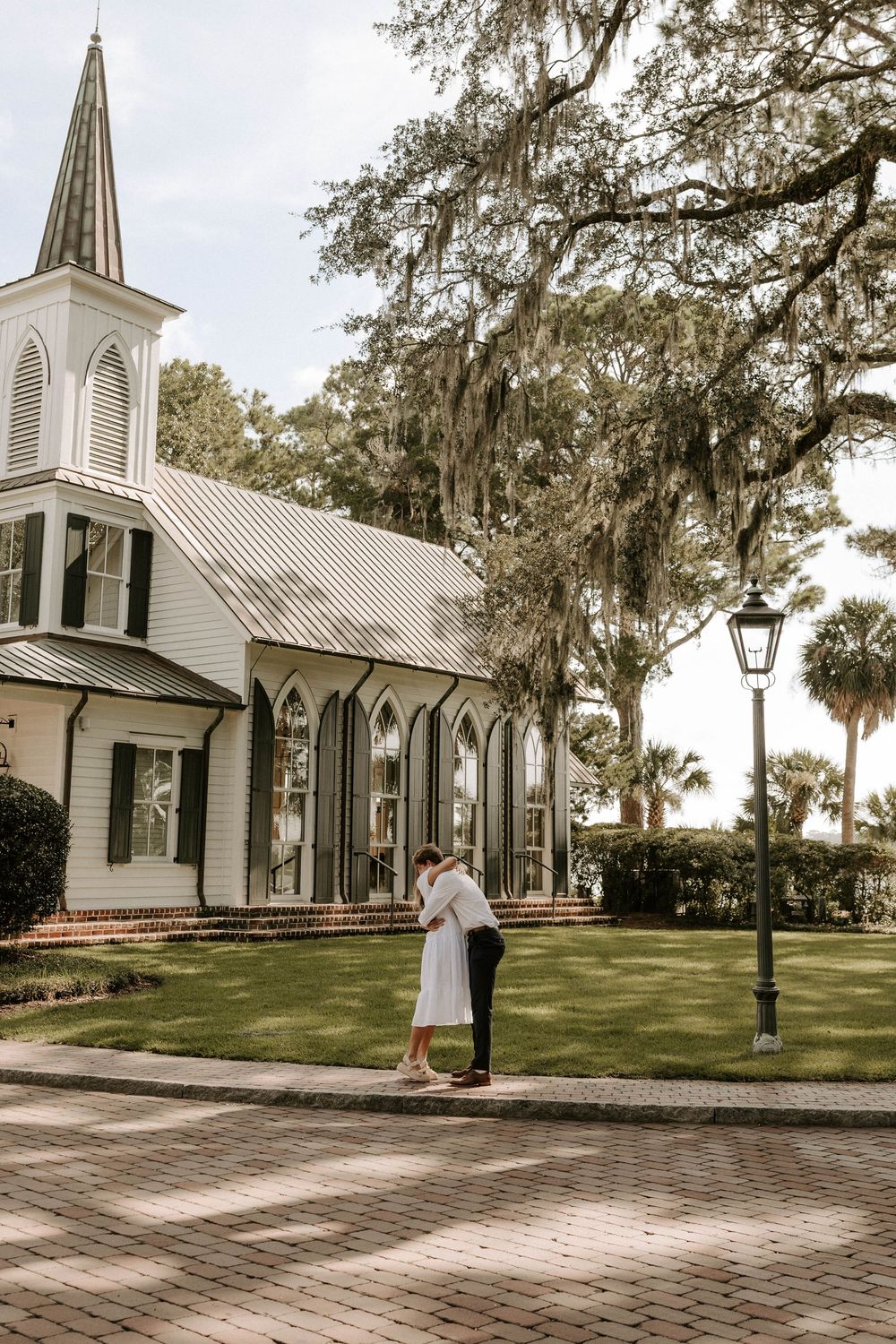 Couple sharing a romantic embrace on a brick path outside a white chapel with Spanish moss trees.