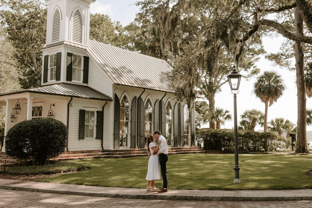 Couple sharing a romantic embrace in front of a charming white chapel surrounded by lush trees.