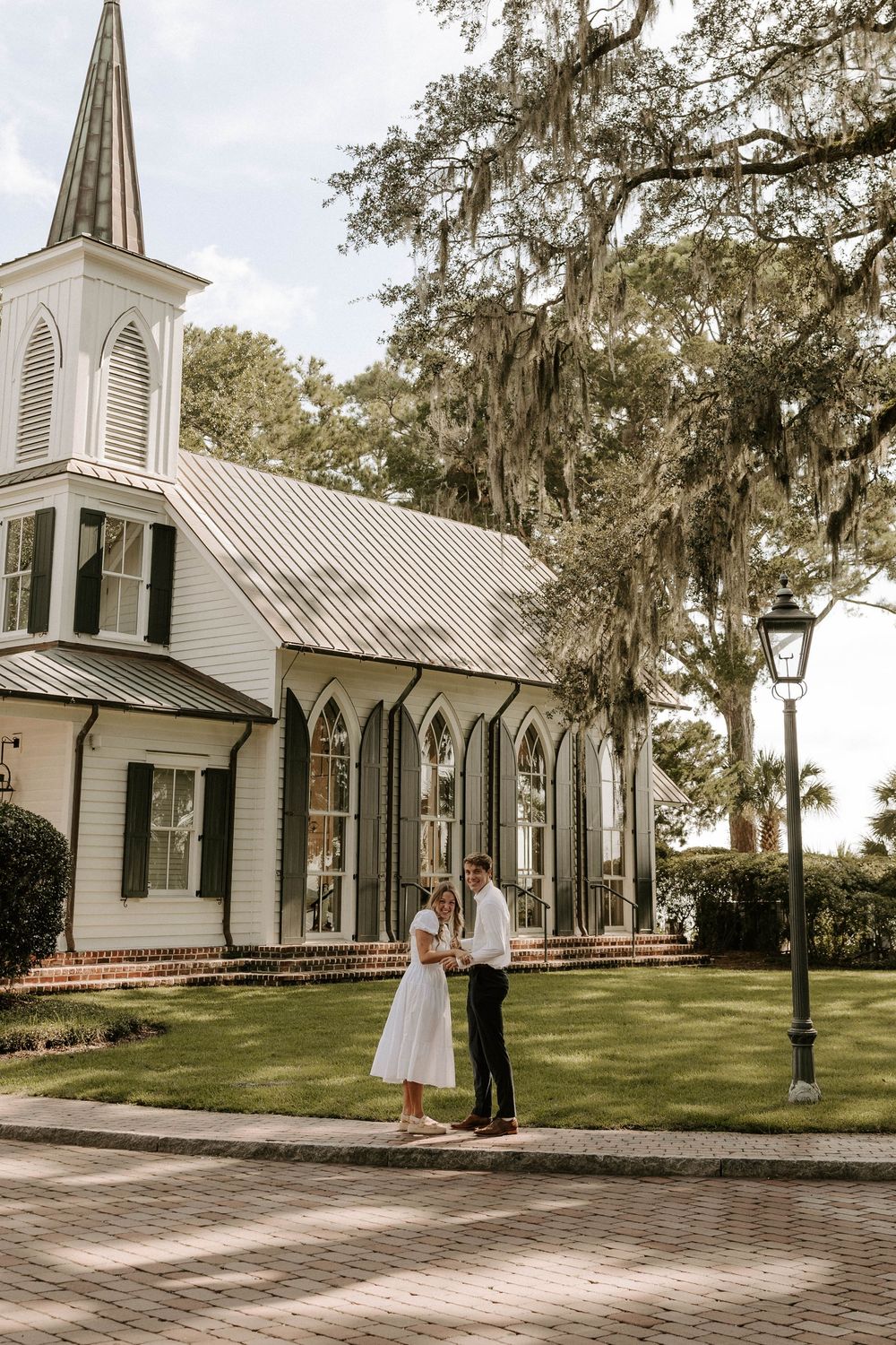 Couple holding hands outside a white Southern chapel surrounded by oak trees draped in Spanish moss.