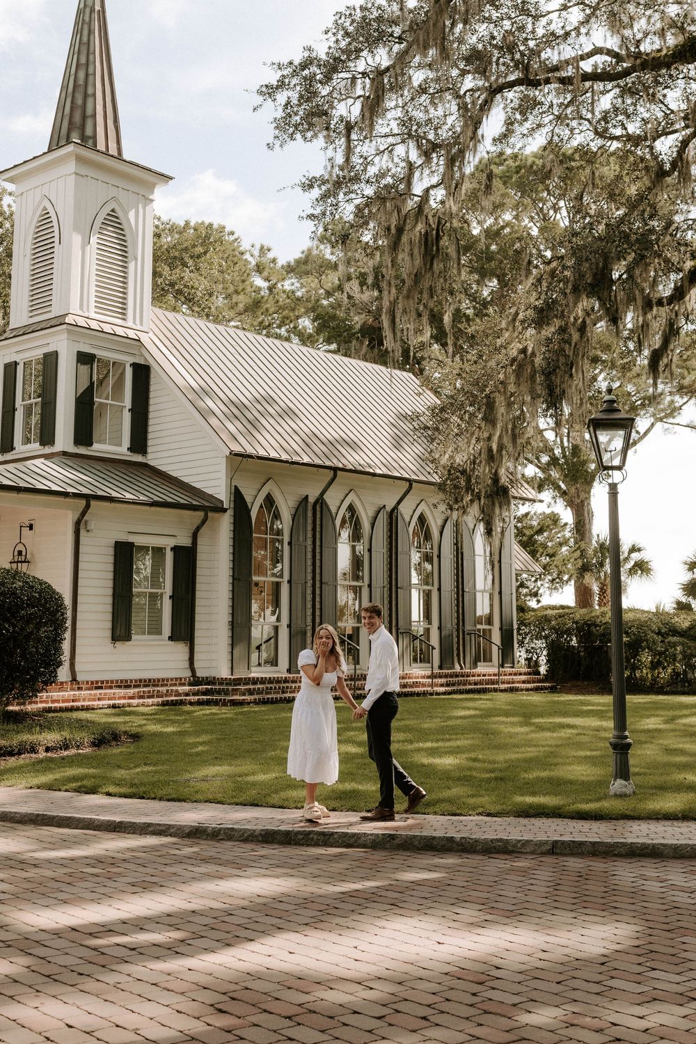 Bride and groom walking hand in hand outside a charming white Southern chapel with gothic windows.