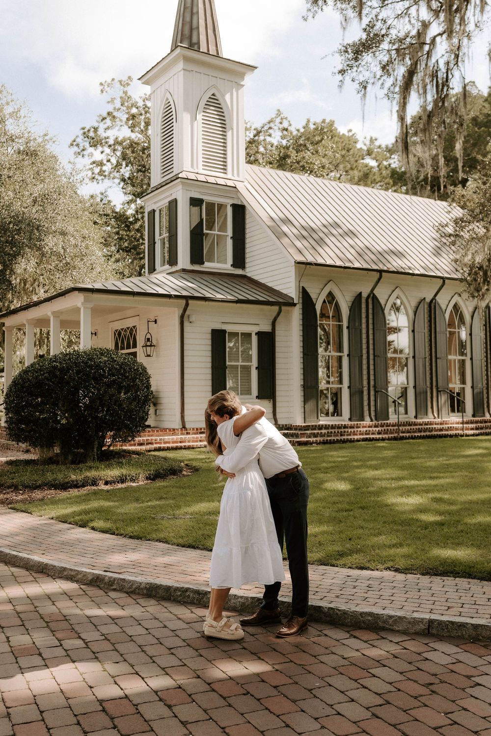 Couple sharing a romantic kiss on brick pathway in front of a white chapel with steeple and gothic windows.