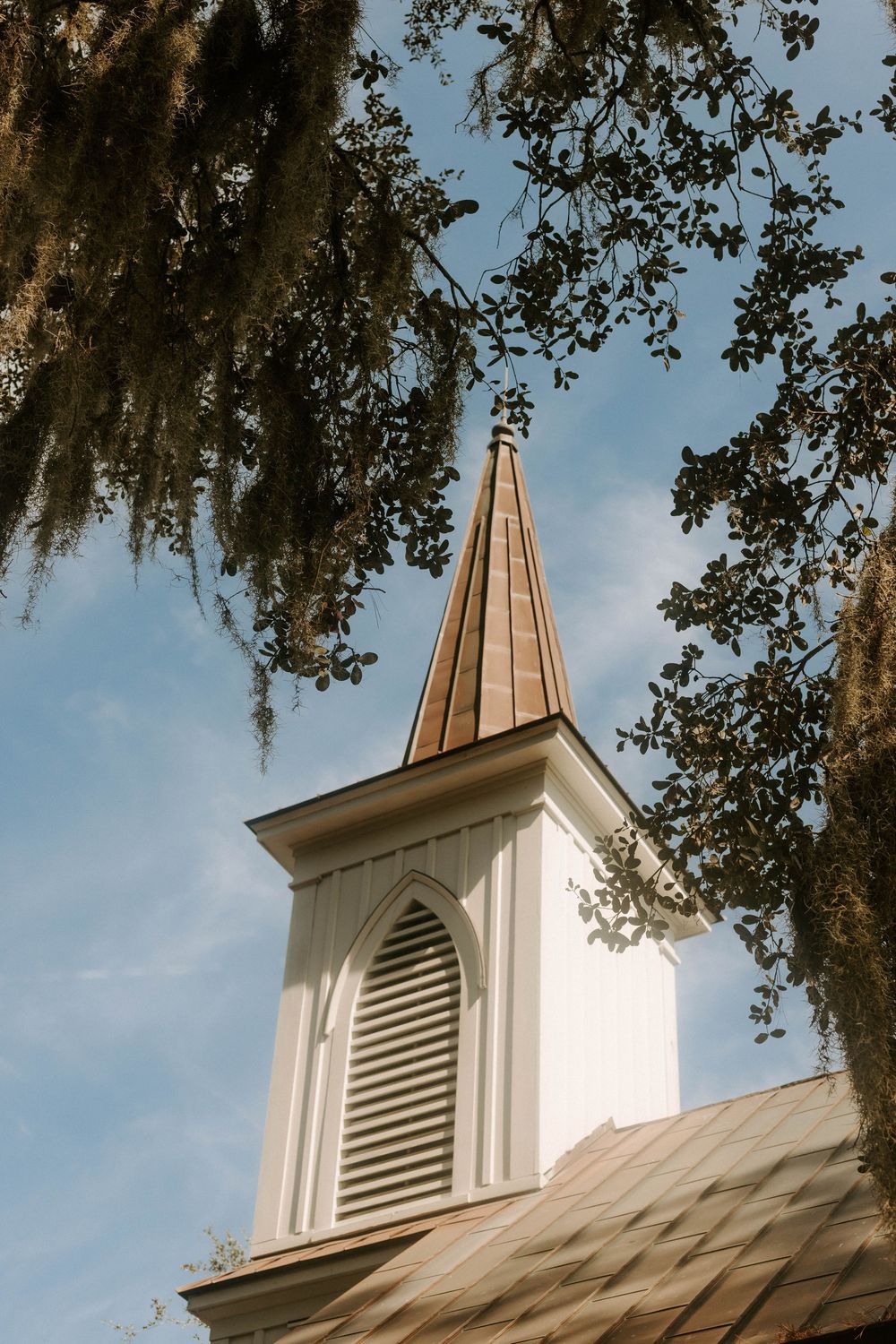White church steeple with copper roof framed by Spanish moss-draped oak trees against a blue sky.