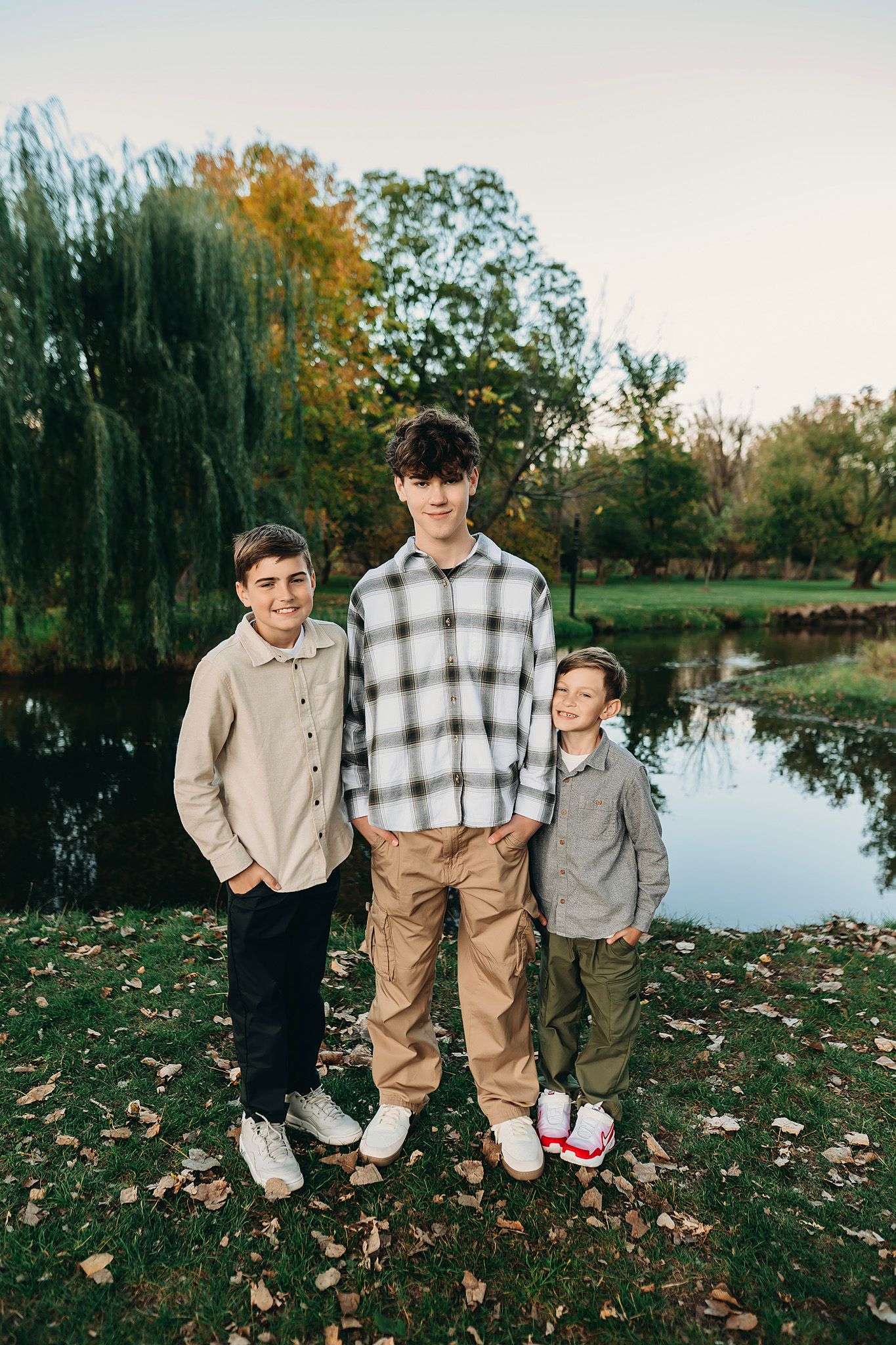 Three boys posing together outdoors near a pond surrounded by autumn trees and fallen