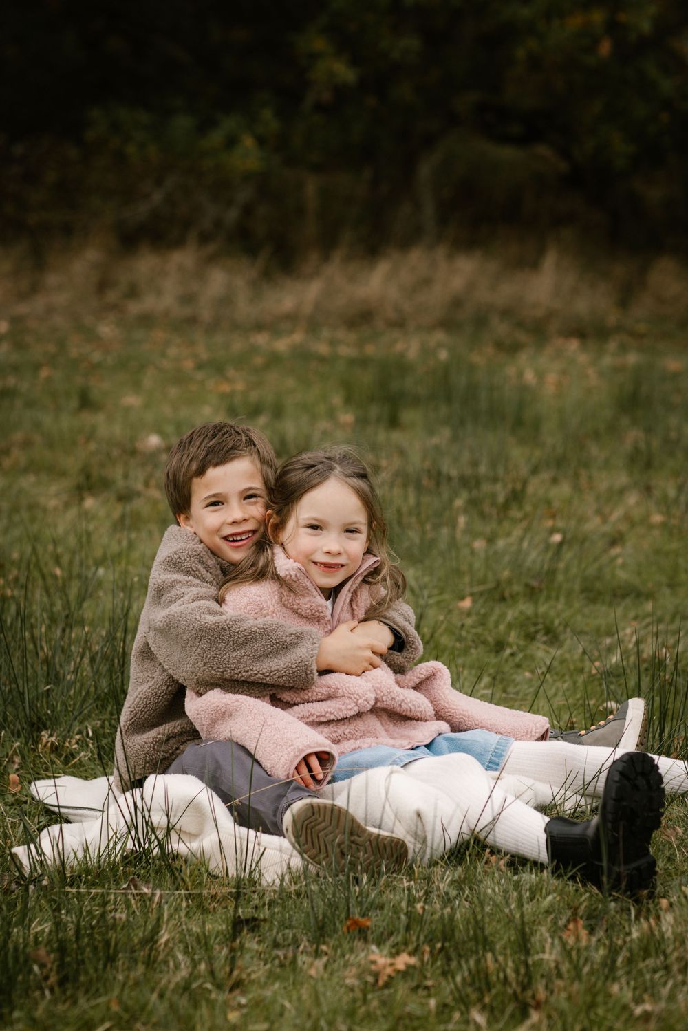 Two young siblings hugging and smiling on a blanket in a grassy autumn field.
