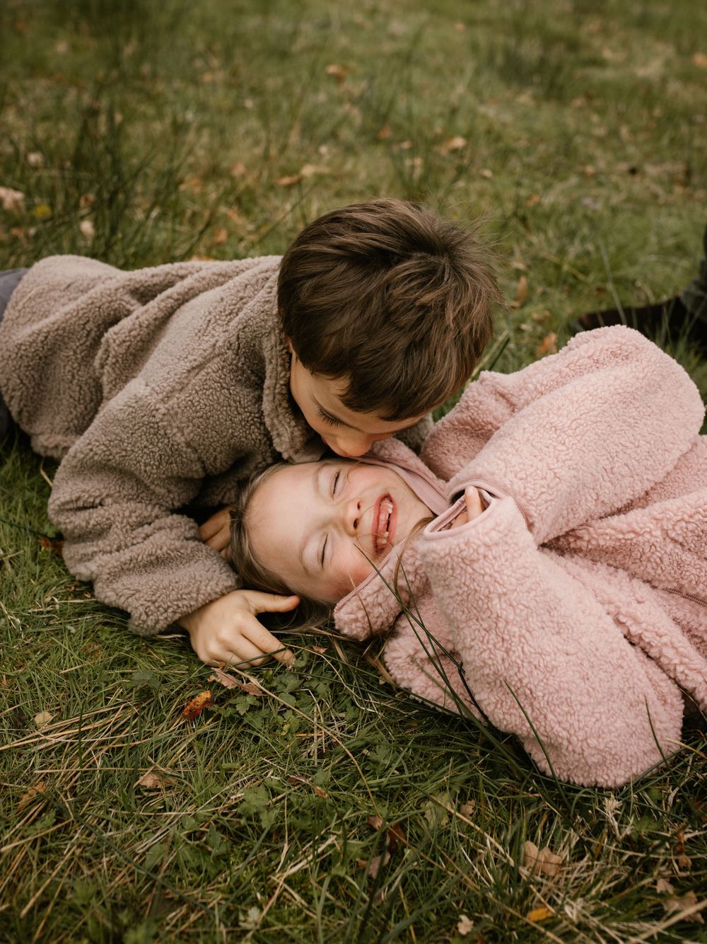 Two children laughing and cuddling on grass in matching teddy fleece coats during an outdoor autumn photo session.