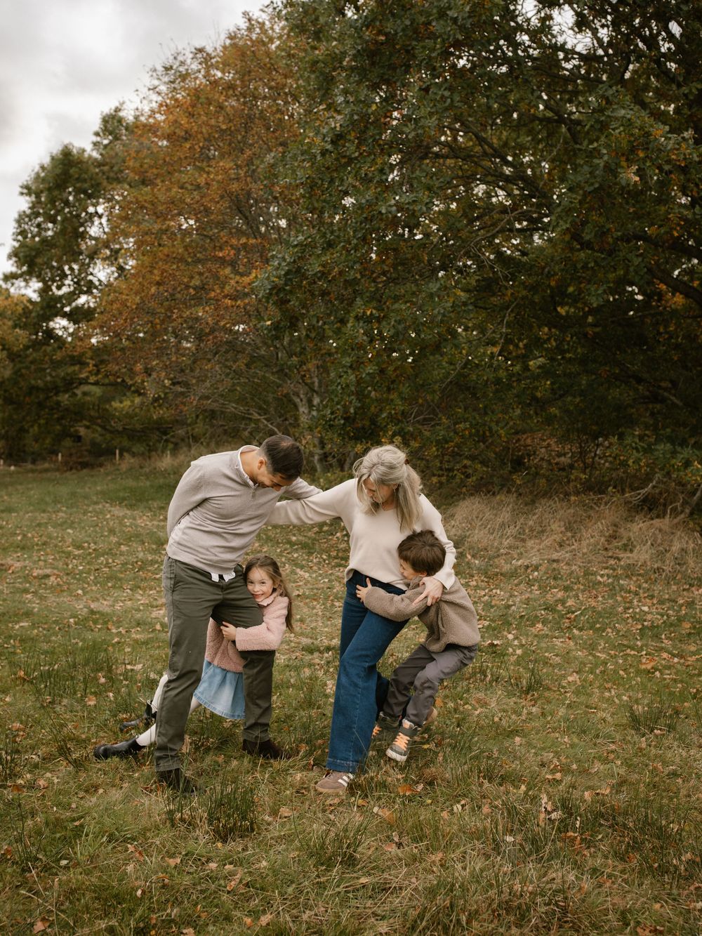 A family of four playfully interacting in a scenic autumn field surrounded by colorful fall foliage.
