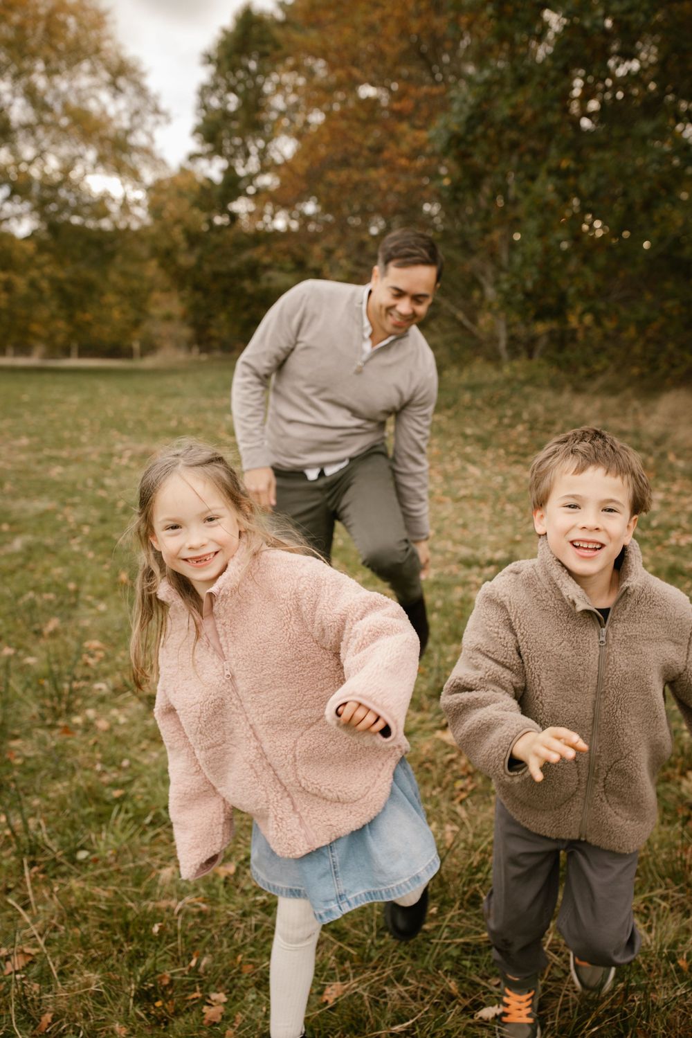 A father chases two laughing children through an autumn park, all wearing cozy sweaters.