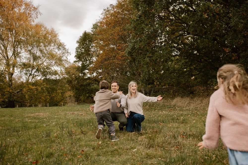 Family playing in autumn field, parents crouching to embrace running child with colorful fall trees in background.
