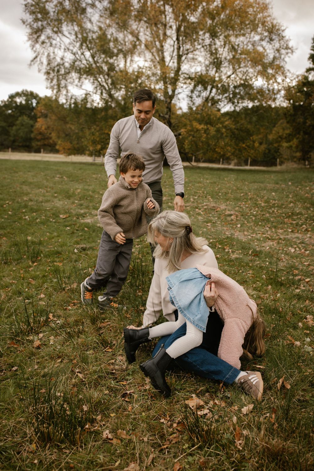 Family playing outdoors in autumn park, father lifting toddler son while mother kneels with young daughter on grass.