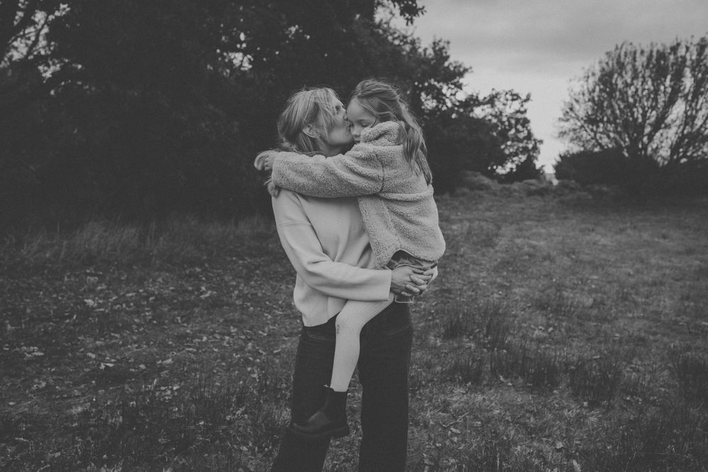 Black and white photo of a woman holding and kissing a young child in a field with trees in the background.