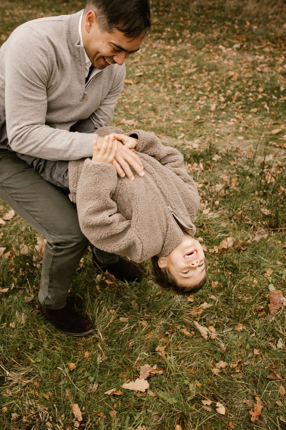 Father playfully holding child upside down on autumn grass, both laughing outdoors in matching beige sweaters.