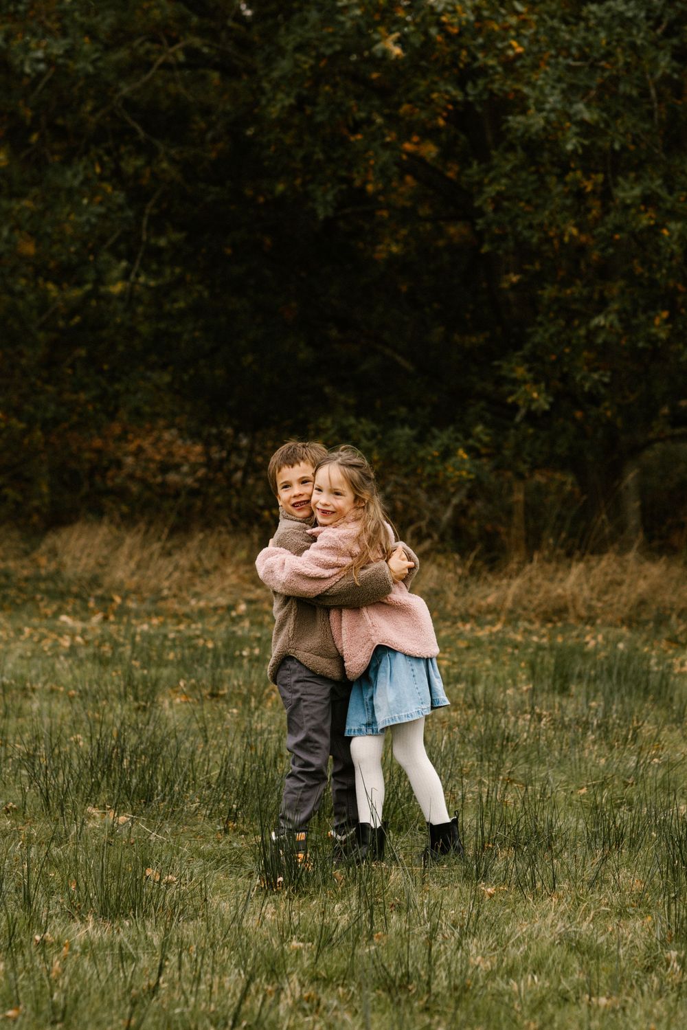 Two young children hugging and smiling in an autumn field with lush green trees in the background.