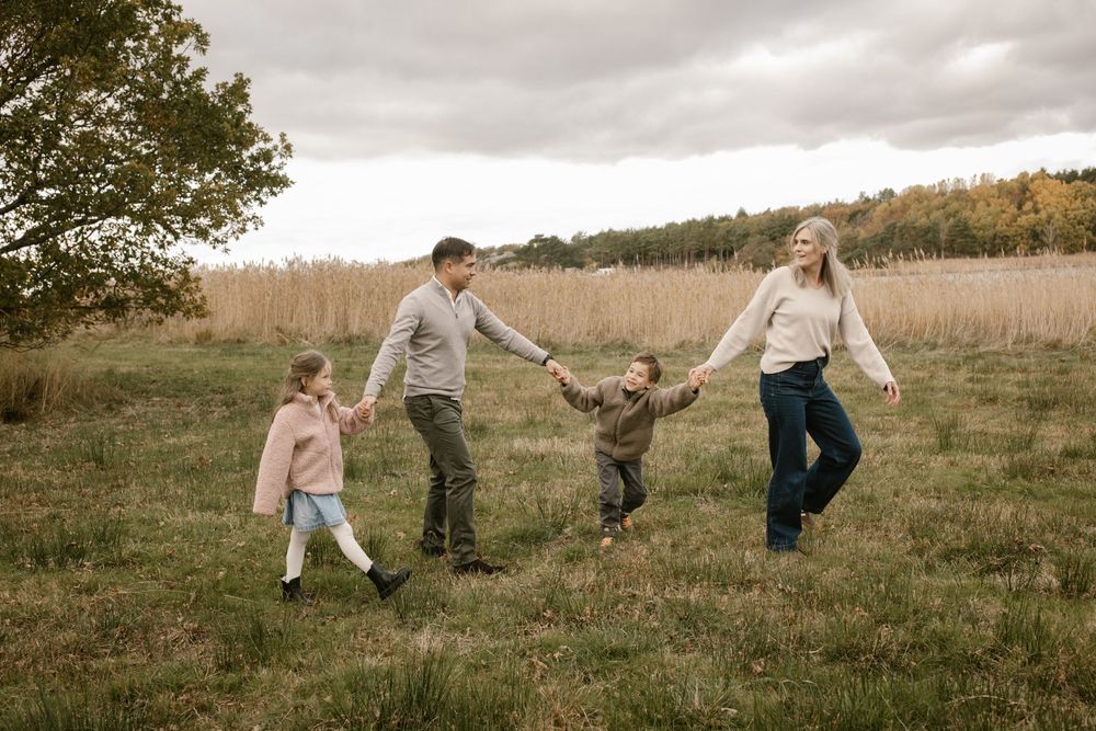Family of four holding hands and playing in an autumn field with colorful trees in the background.