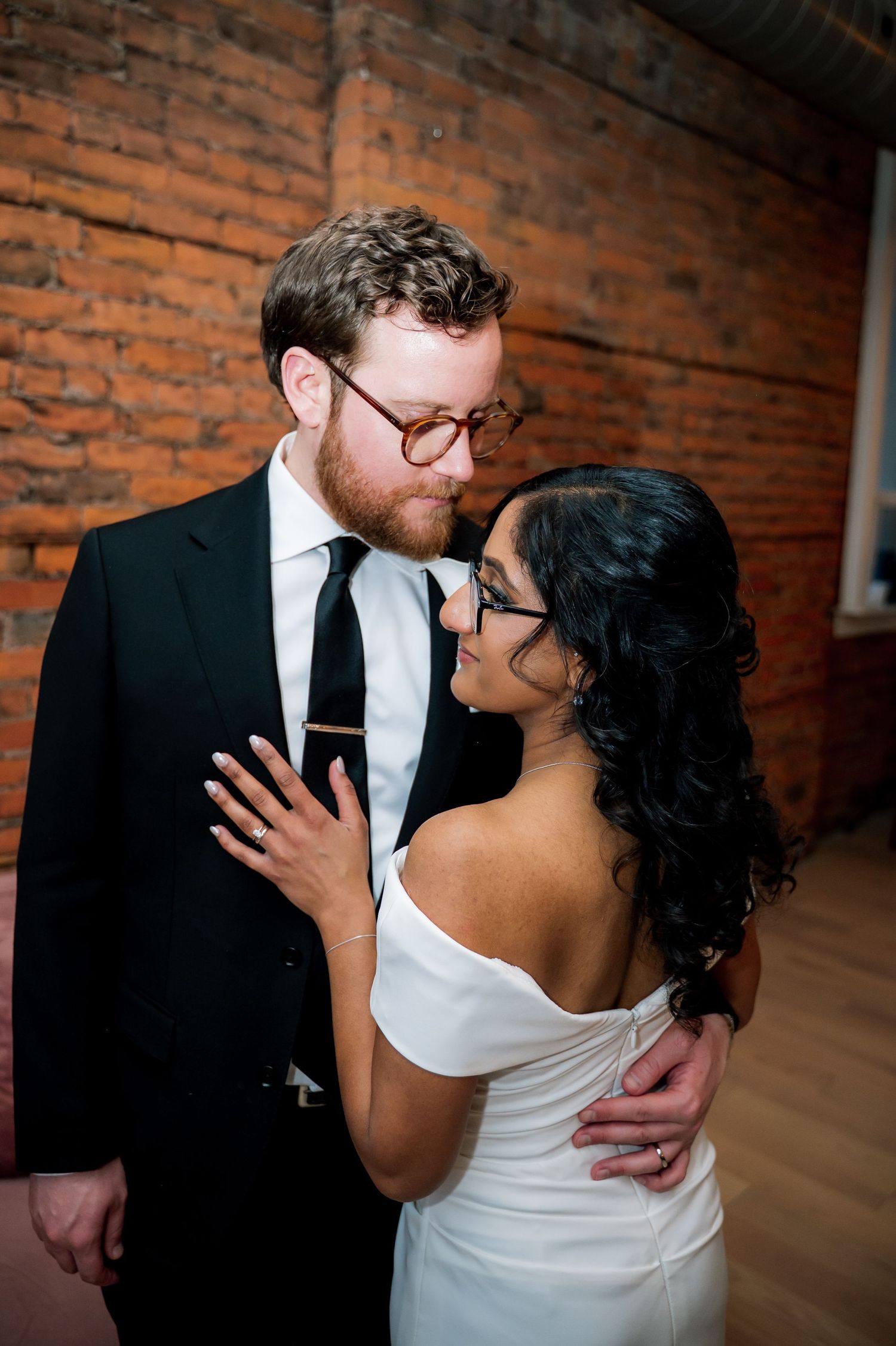 A bride and groom share a romantic moment, she in a white off-shoulder gown, he in a black suit, against a brick wall.