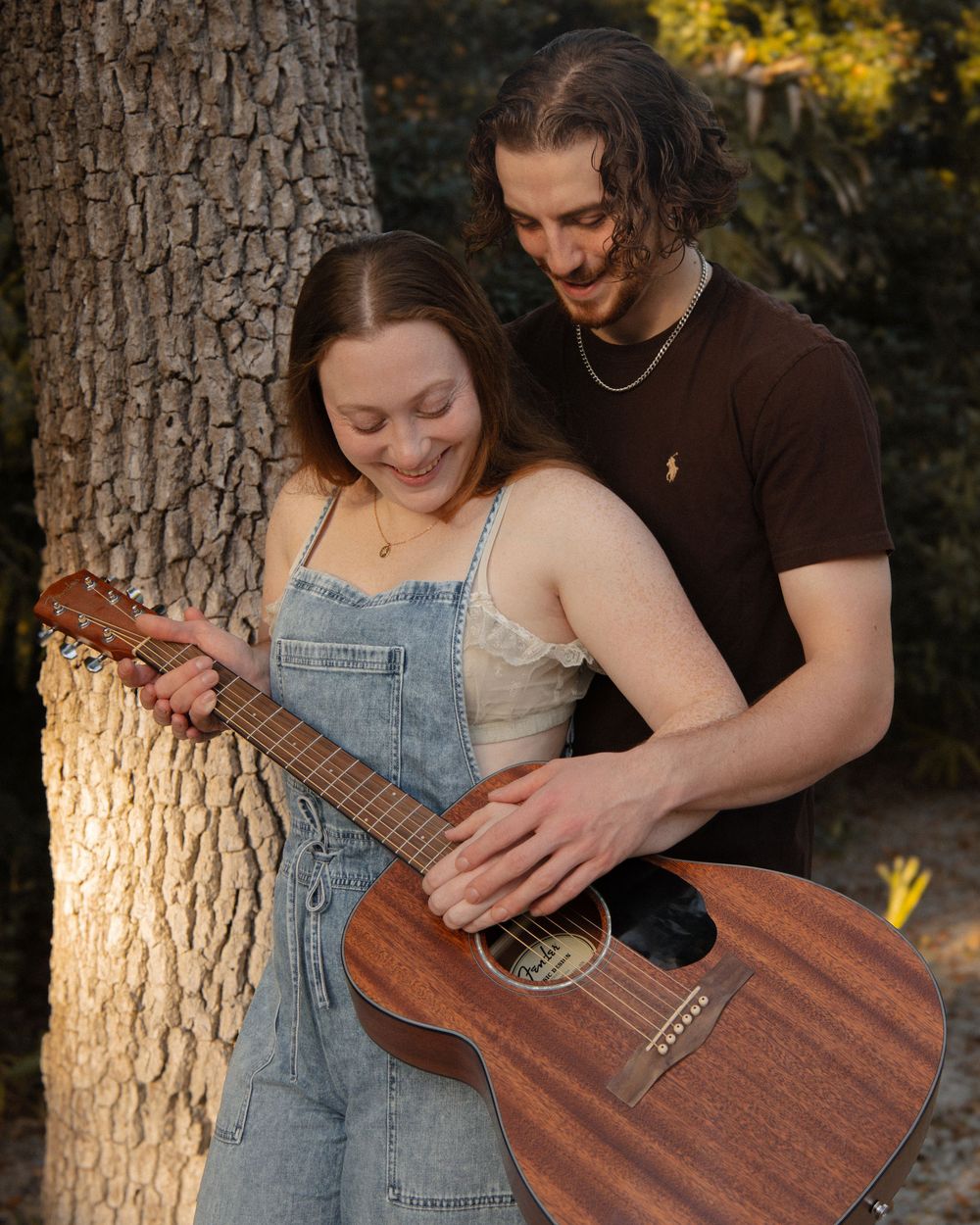 Young couple sharing a tender moment with an acoustic guitar near a tree in golden light.