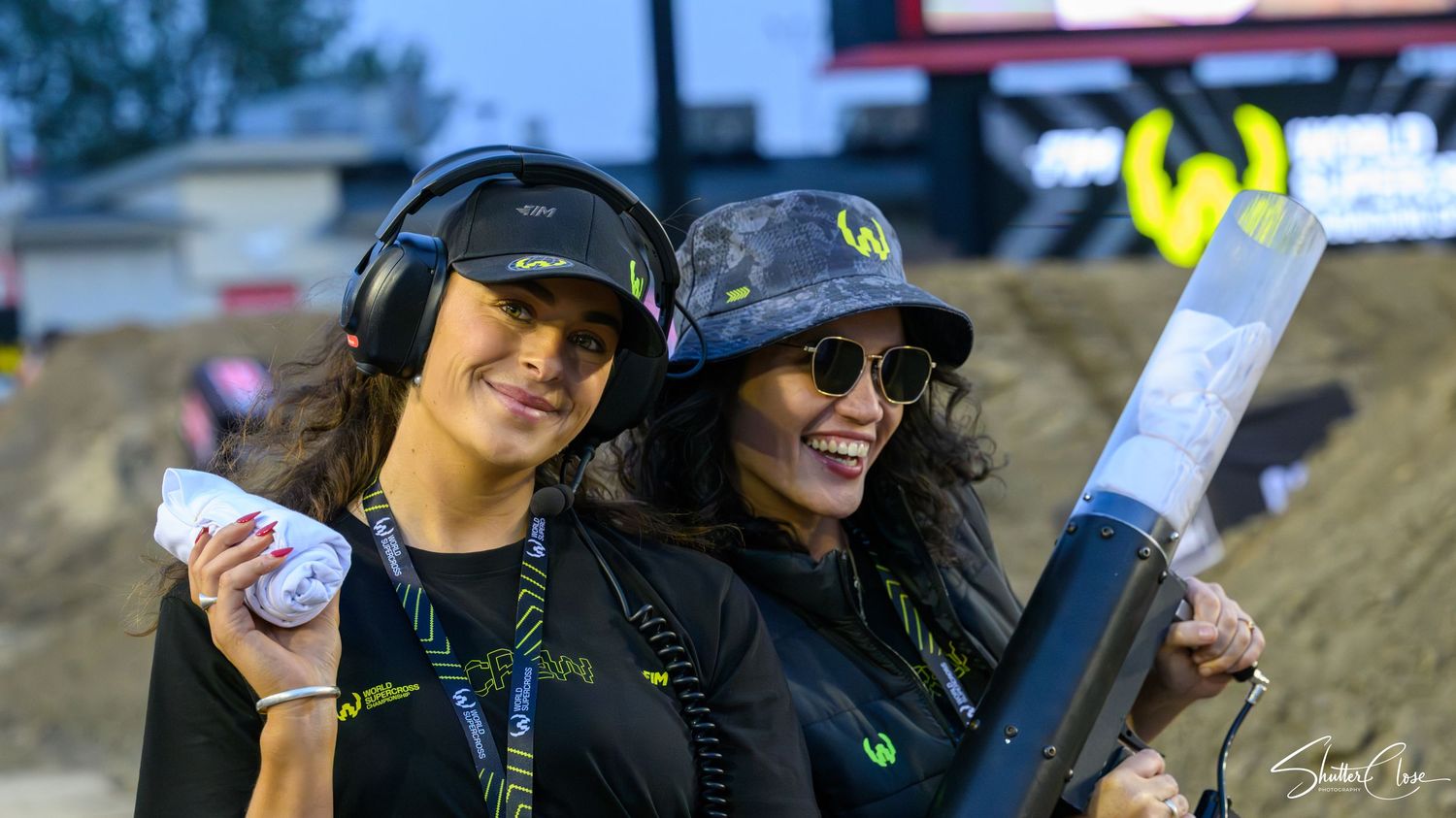 Two women wearing headphones and sunglasses smile at an outdoor motorsport event, one holding a rolled document.