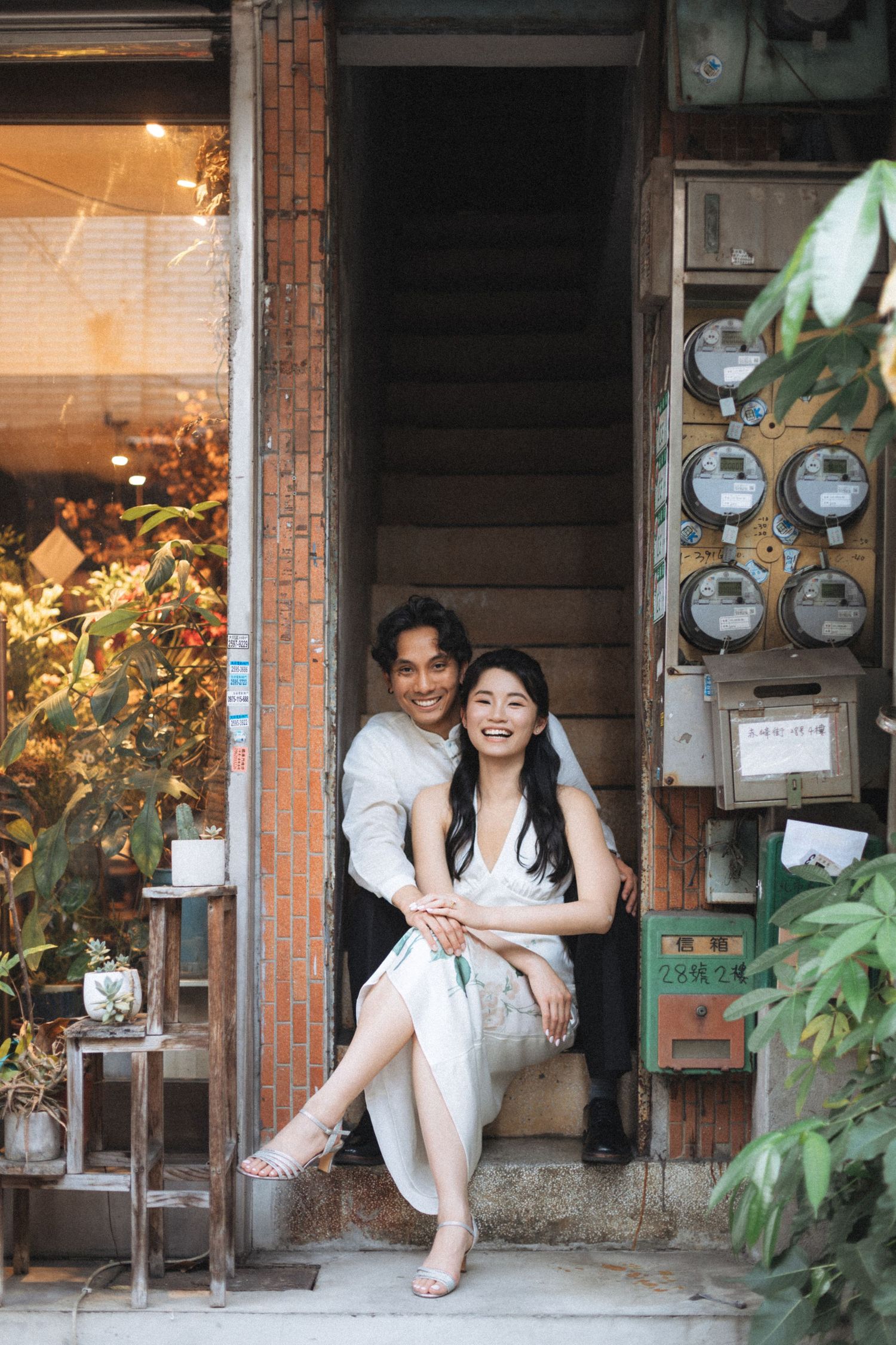 A smiling couple in white outfits sitting on vintage shop steps surrounded by plants and old utility meters.