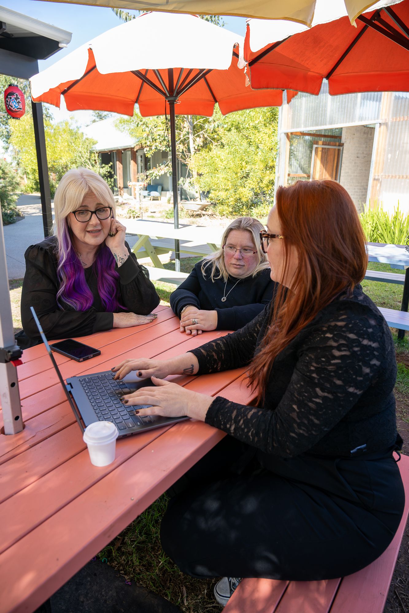 Three women working on a laptop at an outdoor red picnic table under orange and white umbrellas.