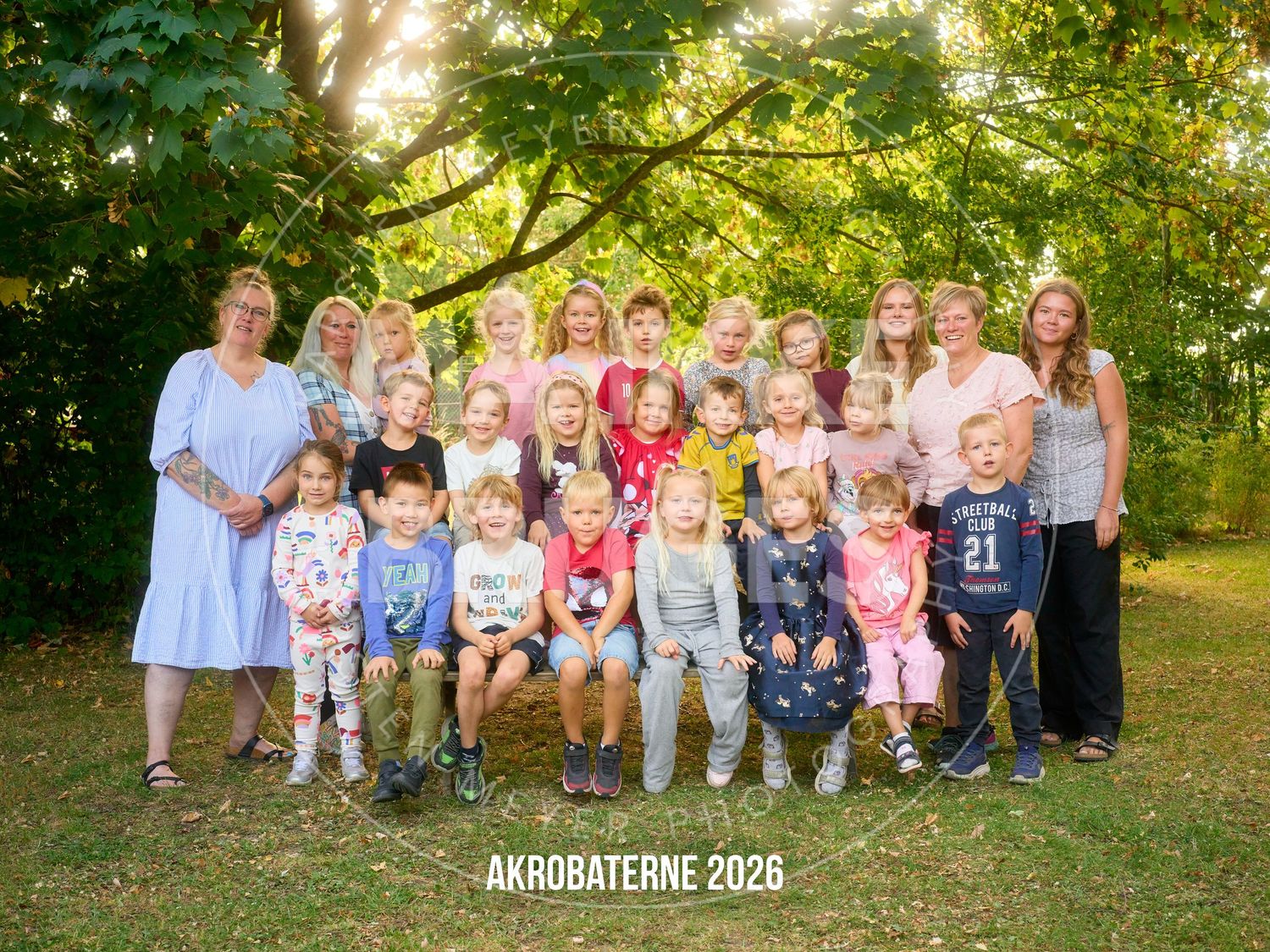 Group photo of Akrobaterne 2026 children and teachers outdoors under green trees in summer.
