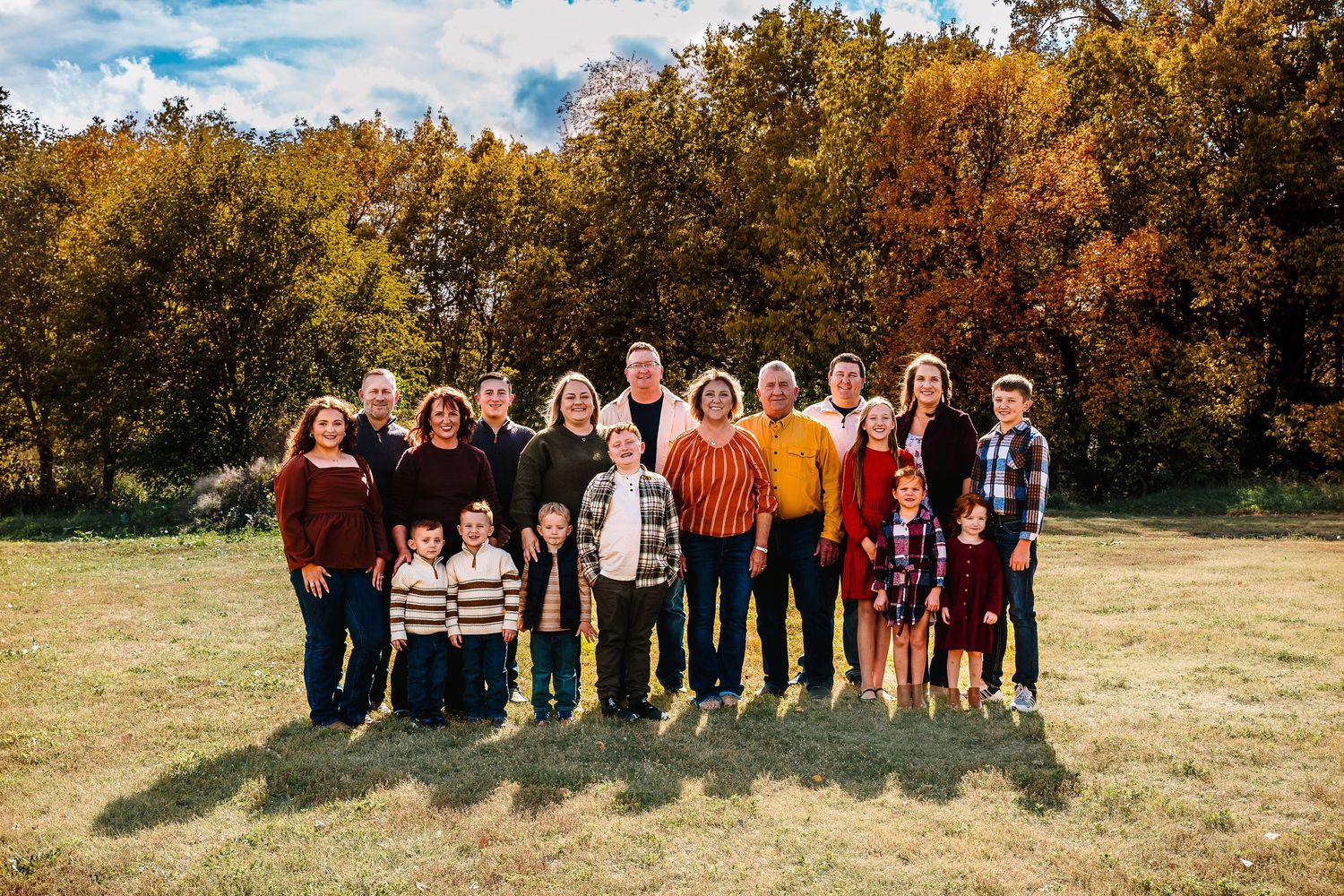 Large family group portrait outdoors in autumn, posing on green grass with colorful fall foliage trees in background.