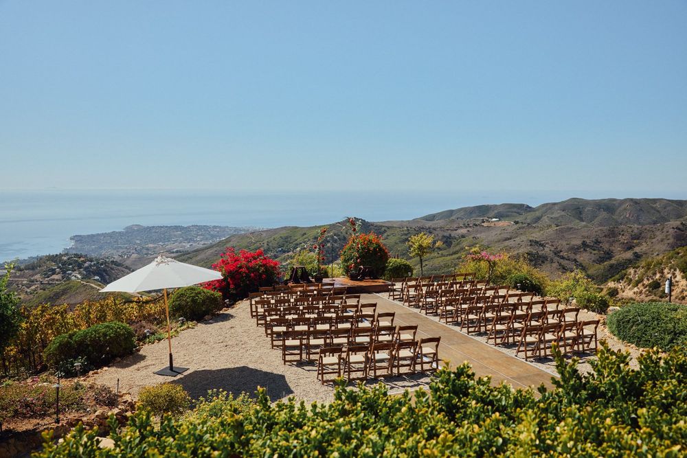 Outdoor wedding ceremony setup with wooden chairs on a hilltop overlooking the Pacific Ocean and coastal hills.