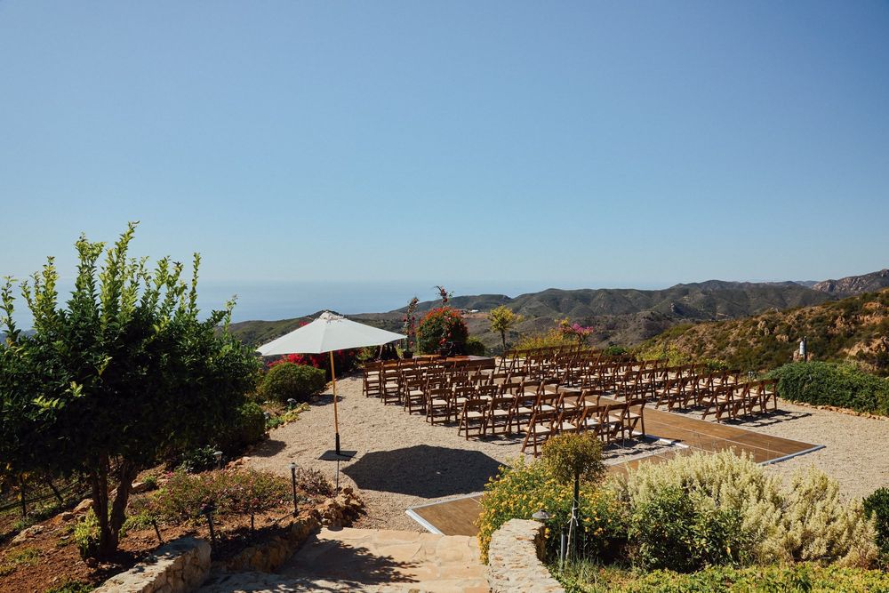 Outdoor wedding ceremony setup with wooden chairs on a hilltop overlooking scenic mountains and ocean under clear blue sky.