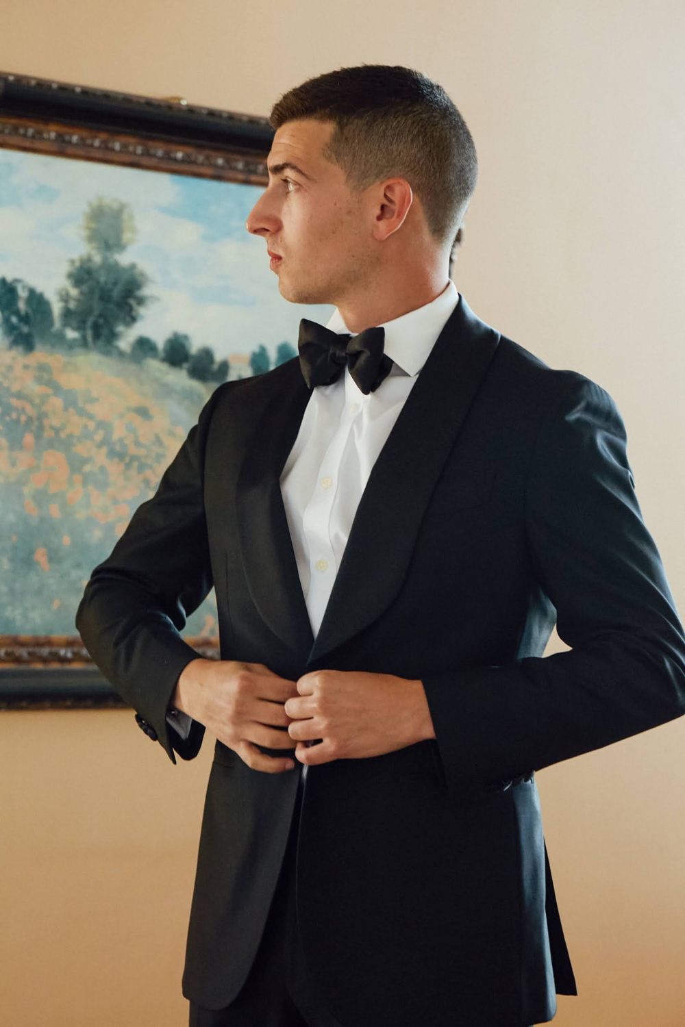 Young man in black tuxedo with bow tie buttoning jacket, standing before impressionist landscape painting.