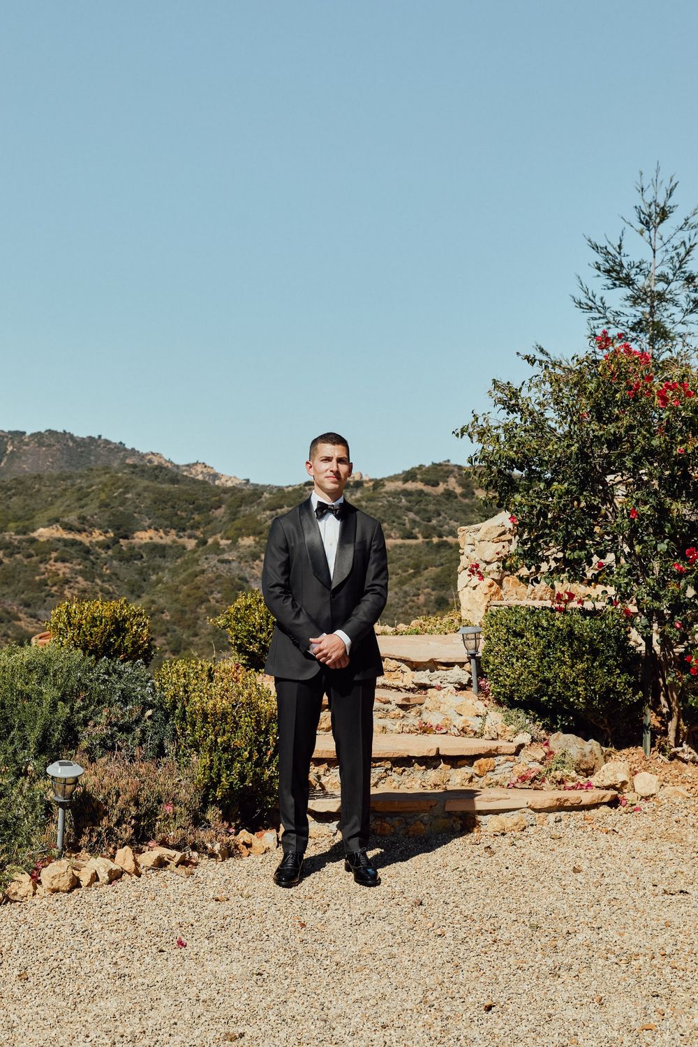 A groom in a black tuxedo stands on a gravel path surrounded by lush greenery and mountains under a clear blue sky.