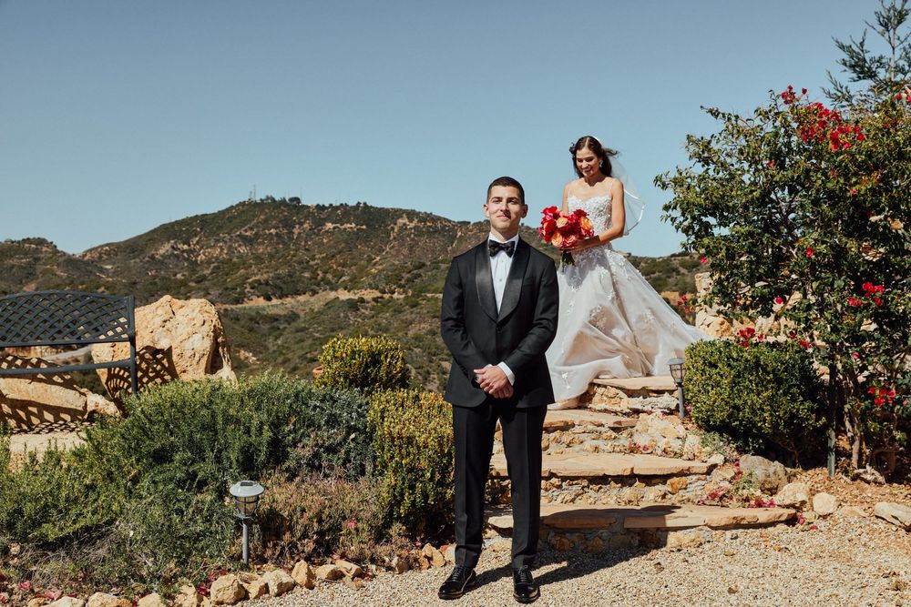 Bride and groom in tuxedo posing outdoors with scenic mountain backdrop and red floral bouquet.