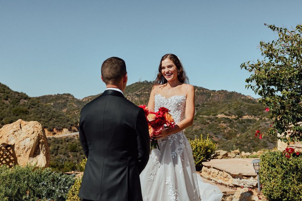 Smiling bride in lace gown holds red bouquet facing groom in black suit during outdoor wedding ceremony.
