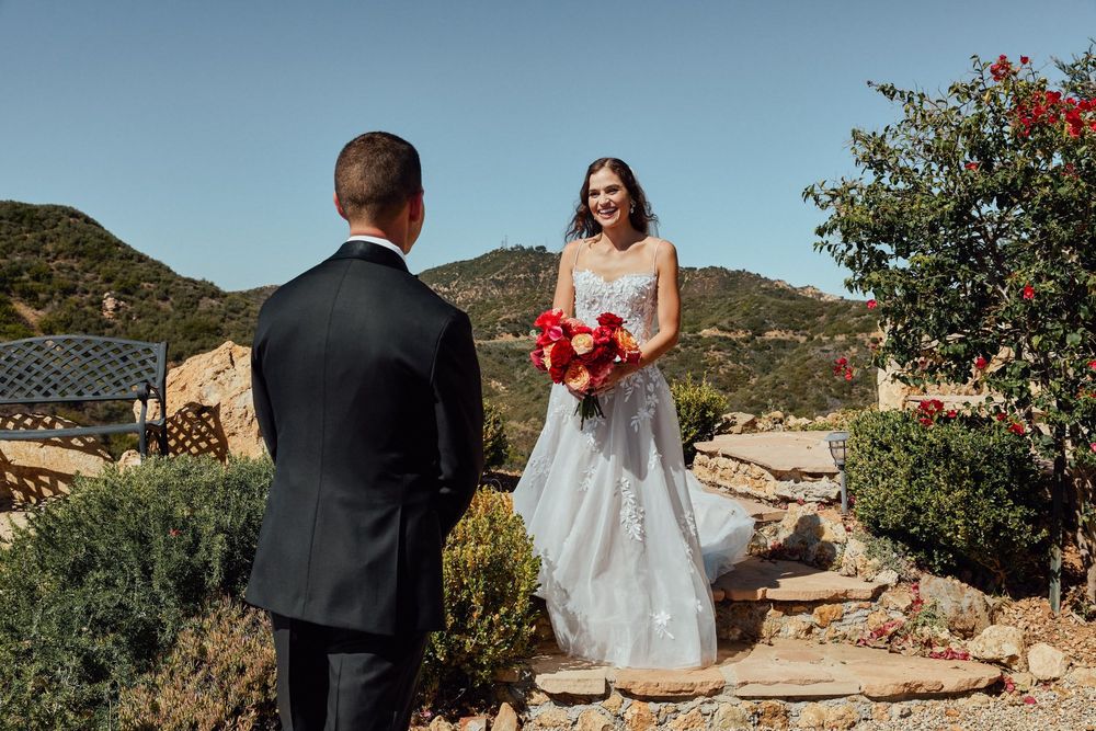 Bride in white lace gown holds red bouquet facing groom in black suit at outdoor wedding venue with hills.