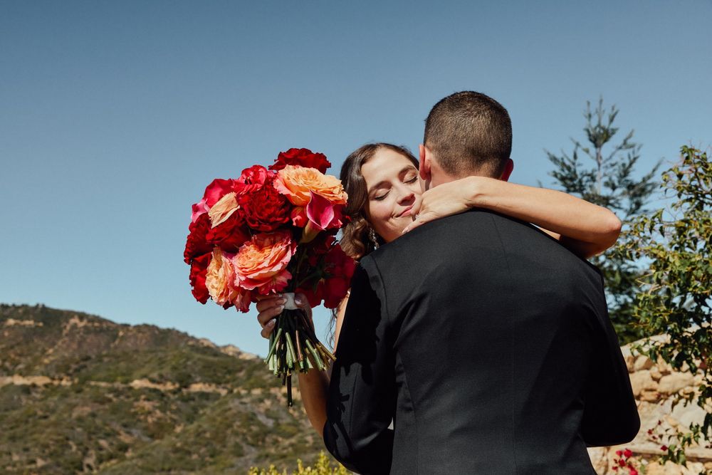 Bride embraces groom while holding vibrant red and orange rose bouquet with scenic mountain backdrop.
